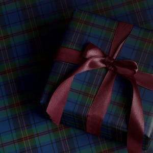 May include: A wrapped gift box with a burgundy ribbon and bow, set on a matching plaid wrapping paper. The plaid pattern features shades of blue, green, and yellow. The gift box is square-shaped.