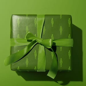 Op de afbeelding: Een groene geschenkdoos verpakt in papier met een patroon van notenkrakerfiguren. Een bijpassend groen lint is in een strik gebonden. De presentatie is feestelijk en decoratief.