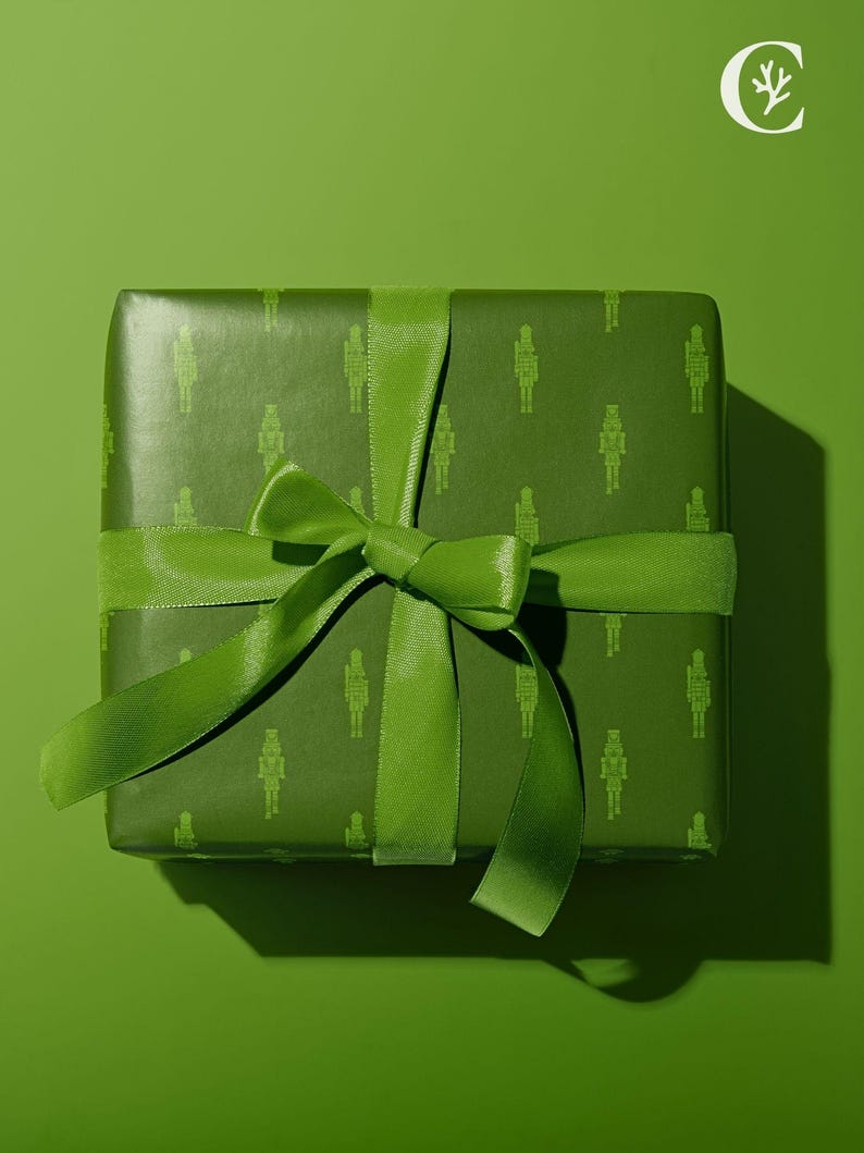 Op de afbeelding: Een groene geschenkdoos verpakt in papier met een patroon van notenkrakerfiguren. Een bijpassend groen lint is in een strik gebonden. De presentatie is feestelijk en decoratief.