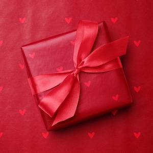 May include: A red gift box wrapped in matching paper and tied with a satin ribbon. The wrapping paper features a pattern of small red hearts. The gift is presented on a red surface with a heart pattern.