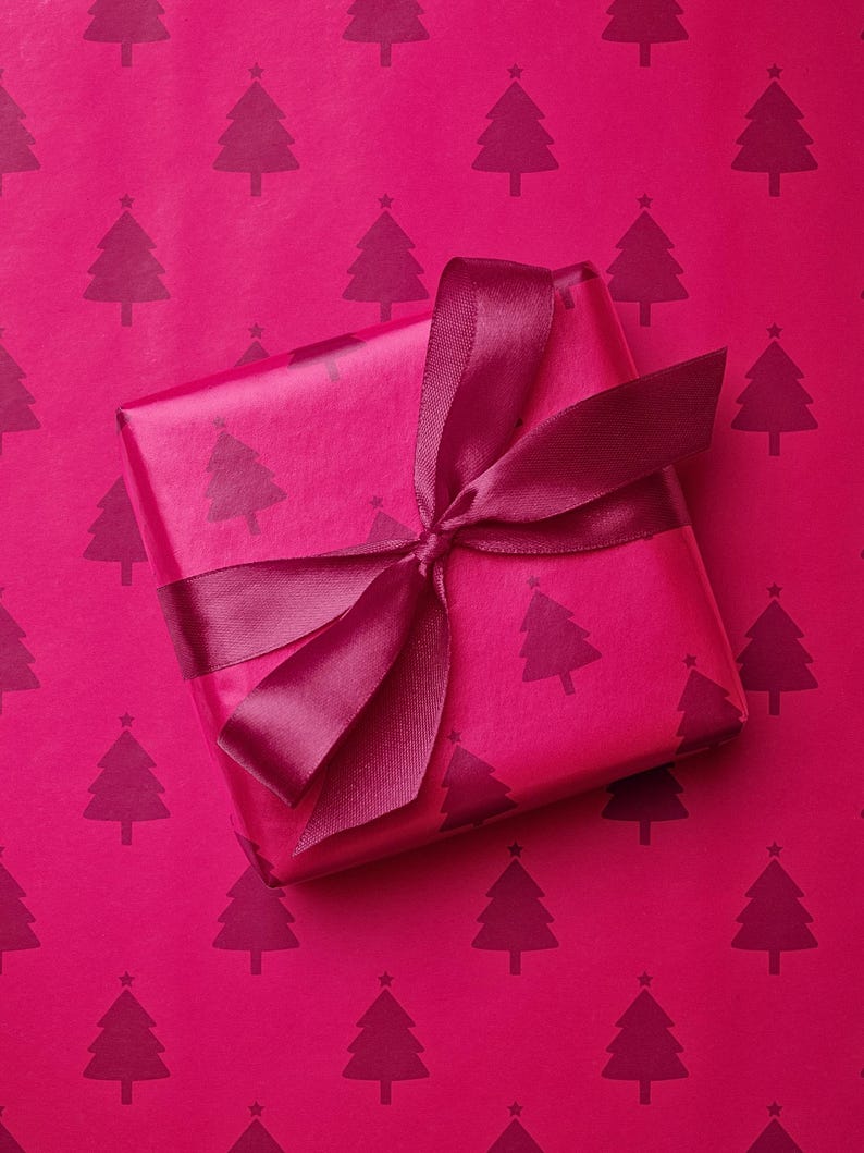 Pu&ograve; includere: Un pacco regalo avvolto su uno sfondo magenta. La carta da regalo e lo sfondo presentano un motivo ripetuto di alberi di Natale magenta scuro. Un nastro magenta scuro &egrave; legato attorno al regalo.