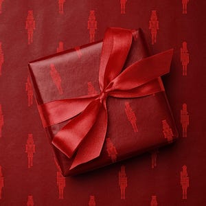 May include: A wrapped gift with a red satin ribbon bow. The gift is wrapped in red paper with a repeating pattern of red nutcracker figures. The background is also red with the same nutcracker pattern.