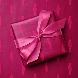 May include: A wrapped gift with a magenta background. The gift is wrapped in magenta paper with a repeating pattern of nutcracker figures. A magenta satin ribbon is tied in a bow on top of the gift.