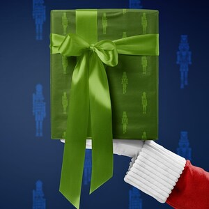 Op de afbeelding: Een ingepakte geschenkdoos met een groen notenkrakerpatroon en een bijpassend groen satijnen lint en strik. Het cadeau wordt vastgehouden door een persoon die een kerstman kostuum draagt, met een blauwe achtergrond.