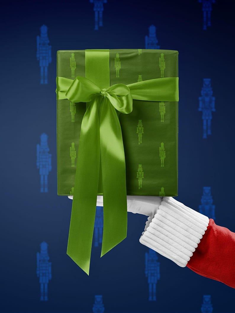 Op de afbeelding: Een ingepakte geschenkdoos met een groen notenkrakerpatroon en een bijpassend groen satijnen lint en strik. Het cadeau wordt vastgehouden door een persoon die een kerstman kostuum draagt, met een blauwe achtergrond.
