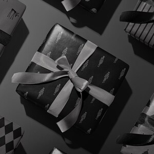 Peut inclure: Vue a&eacute;rienne de plusieurs cadeaux emball&eacute;s avec des rubans et des n&oelig;uds gris. Les cadeaux sont envelopp&eacute;s dans du papier noir avec divers motifs, notamment des flocons de neige, des rennes et le P&egrave;re No&euml;l. Le fond est gris fonc&eacute;.