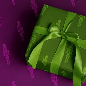 Op de afbeelding: Een ingepakt cadeau met een groene strik en lint op een paarse achtergrond. Het cadeaupapier is groen met een herhaald patroon van notenkrakersoldaten. De achtergrond heeft een herhaald patroon van roze notenkrakersoldaten.