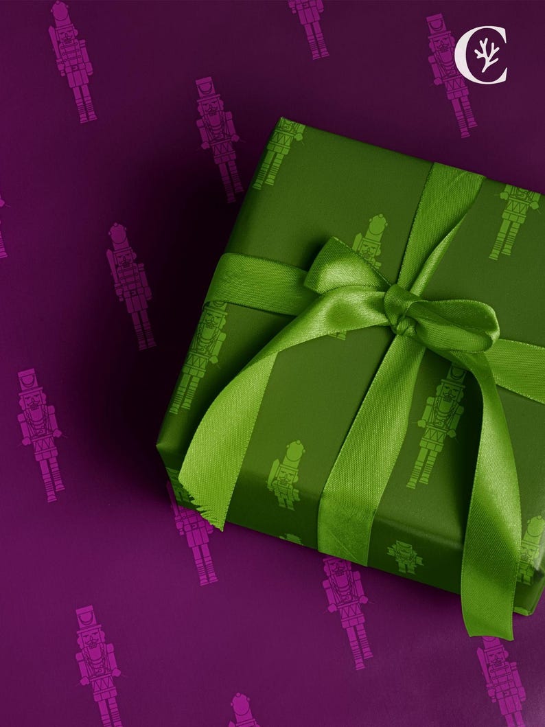 Op de afbeelding: Een ingepakt cadeau met een groene strik en lint op een paarse achtergrond. Het cadeaupapier is groen met een herhaald patroon van notenkrakersoldaten. De achtergrond heeft een herhaald patroon van roze notenkrakersoldaten.