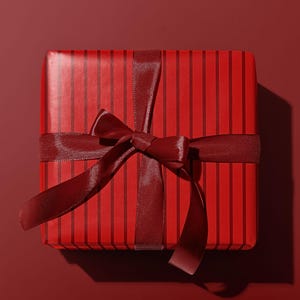 May include: A square gift box wrapped in red paper with vertical dark red stripes. A dark red satin ribbon is tied around the box with a bow on top. The background is a solid red.