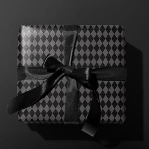 Peut inclure: Un coffret cadeau carr&eacute; envelopp&eacute; dans du papier noir avec un motif de losanges r&eacute;p&eacute;t&eacute;s. Un ruban de satin noir est nou&eacute; autour de la bo&icirc;te en forme de n&oelig;ud. Le fond est noir uni.