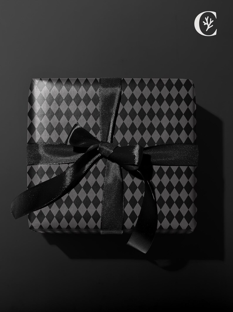 Peut inclure: Un coffret cadeau carr&eacute; envelopp&eacute; dans du papier noir avec un motif de losanges r&eacute;p&eacute;t&eacute;s. Un ruban de satin noir est nou&eacute; autour de la bo&icirc;te en forme de n&oelig;ud. Le fond est noir uni.