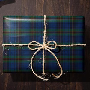May include: A rectangular gift wrapped in blue, green, and brown plaid paper, tied with natural twine. The twine is tied in a bow on top of the package. The background is a dark brown wooden surface.