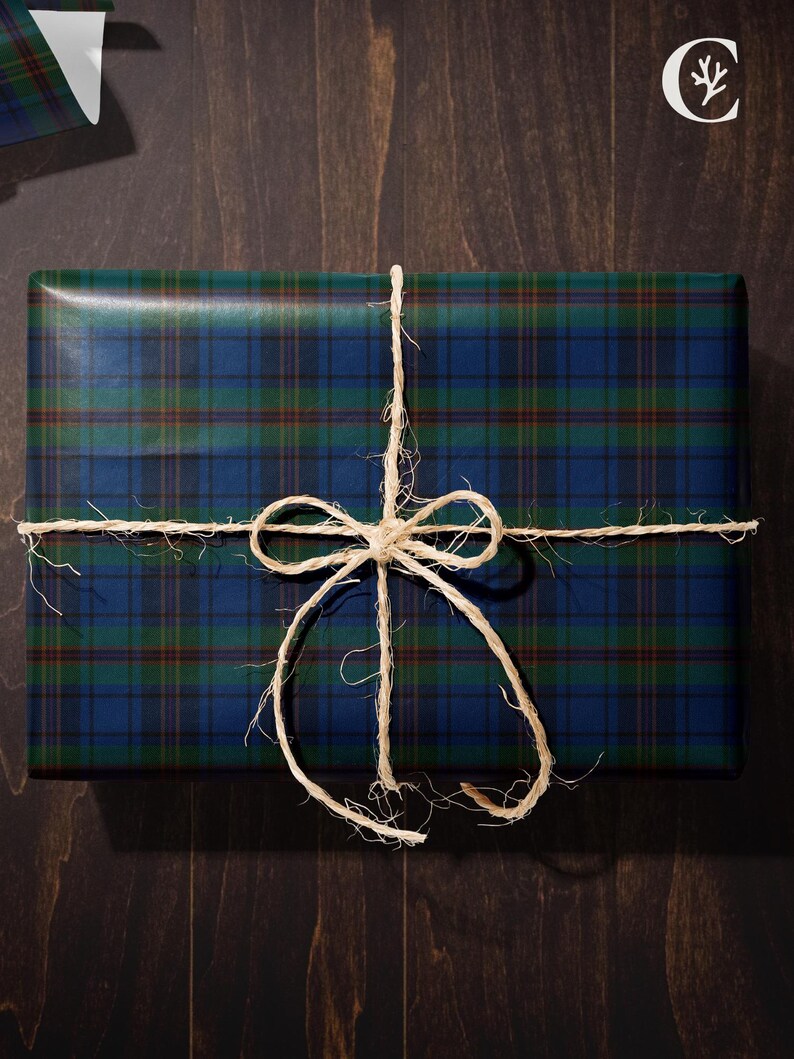 May include: A rectangular gift wrapped in blue, green, and brown plaid paper, tied with natural twine. The twine is tied in a bow on top of the package. The background is a dark brown wooden surface.