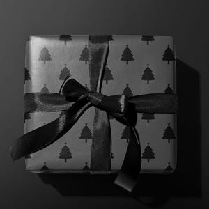 Pode incluir: Uma caixa de presente quadrada embrulhada em papel preto com um padr&atilde;o repetido de &aacute;rvores de Natal. &Eacute; amarrada com uma fita de cetim preta, formando um la&ccedil;o. O fundo &eacute; preto liso.