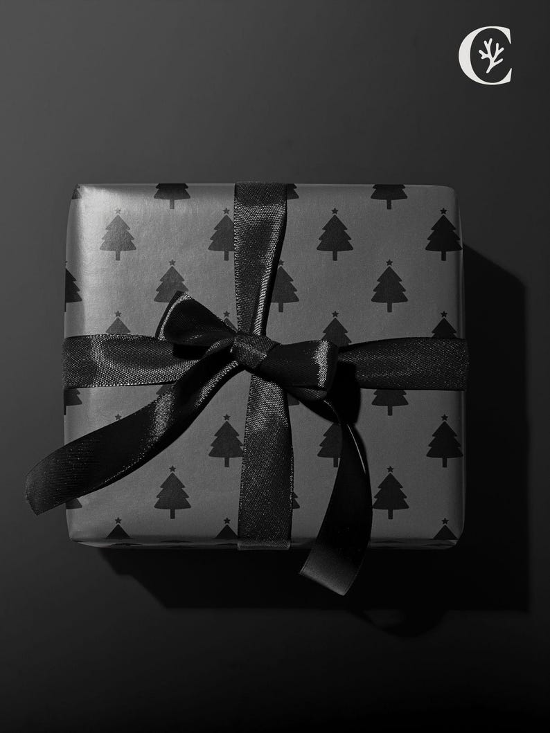 Pode incluir: Uma caixa de presente quadrada embrulhada em papel preto com um padr&atilde;o repetido de &aacute;rvores de Natal. &Eacute; amarrada com uma fita de cetim preta, formando um la&ccedil;o. O fundo &eacute; preto liso.