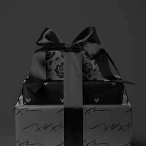 May include: Three stacked gift boxes wrapped in black paper with a large black satin bow. The top box features a rose pattern, the middle box has heart shapes, and the bottom box has the text "xoxo" printed on it.