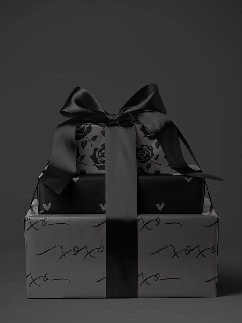 May include: Three stacked gift boxes wrapped in black paper with a large black satin bow. The top box features a rose pattern, the middle box has heart shapes, and the bottom box has the text "xoxo" printed on it.