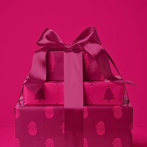 Pu&ograve; includere: Tre scatole regalo impilate, avvolte in carta magenta e legate con un nastro di raso abbinato. Le scatole sono decorate con motivi festivi, tra cui facce di Babbo Natale, alberi di Natale e illustrazioni di soldatini giocattolo. Lo sfondo &egrave; magenta pieno.