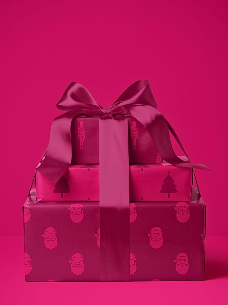 Pu&ograve; includere: Tre scatole regalo impilate, avvolte in carta magenta e legate con un nastro di raso abbinato. Le scatole sono decorate con motivi festivi, tra cui facce di Babbo Natale, alberi di Natale e illustrazioni di soldatini giocattolo. Lo sfondo &egrave; magenta pieno.