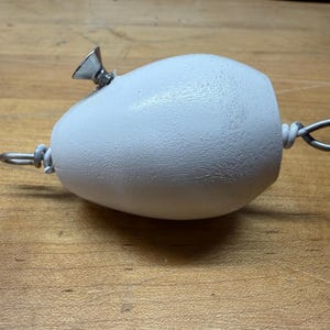 Könnte beinhalten: Ein weißes, eiförmiges Objekt mit einer glatten, matten Oberfläche. Es verfügt über ein kleines, silberfarbenes, kegelförmiges Metallteil an der Oberseite und eine silberne Metalldrahtschlaufe an jedem Ende, die mit verdrilltem Draht befestigt ist.