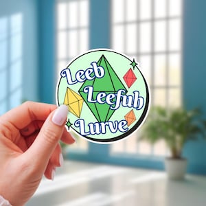 以下が含まれることがあります： 「Leeb Leefub Lurve」という言葉が書かれた丸いステッカー。ステッカーには、緑、黄色、赤、オレンジの幾何学的な形が、黒い縁取りのある淡い緑色の背景に描かれています。ステッカーは手で持たれています。