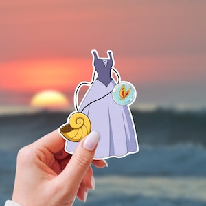 Op de afbeelding: Een sticker met een cartoonillustratie van een lavendelkleurige jurk met een zeeschelp en een vlinderhanger. De jurk heeft een donkerpaars lijfje en een zwierige rok. De zeeschelp is goudkleurig en de vlinder is oranje.