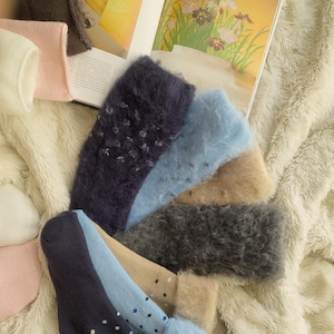 Peut inclure: Une collection de chaussettes douces et duveteuses de différentes couleurs, dont bleu marine, bleu clair, beige et gris. Certaines chaussettes présentent de petits pois blancs ou bleus. Les chaussettes sont disposées sur une surface douce et blanche texturée.