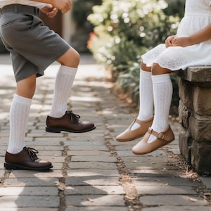 Puede incluir: Dos niños con zapatos clásicos de cuero marrón. Un par es con cordones, el otro es un estilo Mary Jane con hebilla. Ambos niños llevan calcetines altos blancos con un diseño estampado. Un niño lleva pantalones cortos grises.
