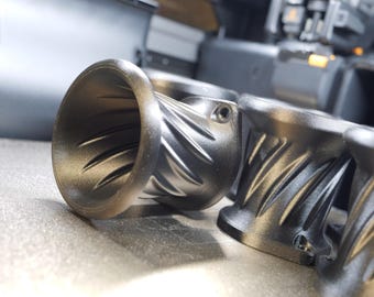 May include: A set of matte black velocity stacks featuring a flared circular opening and a spiraled, ribbed exterior design. Each component includes a small mounting hole on the side flange, showcasing a textured finish and a hollow interior.