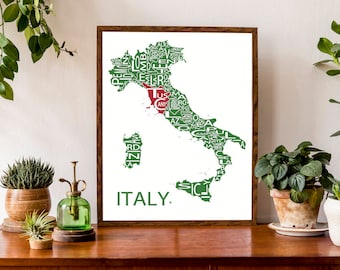 May include: A framed print of a map of Italy with the names of each region written in green and white. The region of Tuscany is highlighted in red. The print is on a white background and has a brown frame. The print is sitting on a wooden table with plants in pots.