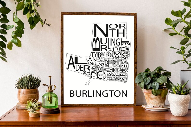 May include: A black and white typography print of the city of Burlington, Vermont. The print features the names of neighborhoods within the city, arranged in the shape of the city's outline. The print is framed in a brown wood frame and is displayed on a wooden table.