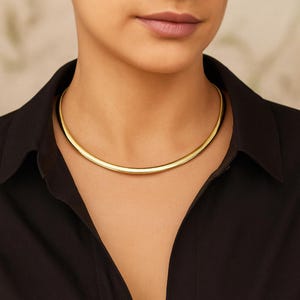 May include: A gold-coloured necklace with a smooth, rounded design. The necklace is worn around the neck and is set against a black shirt. The jewellery has a polished finish, reflecting light. The necklace is a choker style.