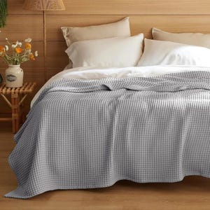 Puede incluir: Una manta gris claro con tejido de gofre sobre una cama. La manta cubre la cama, con almohadas y sábanas blancas visibles. La cama está contra un cabecero de madera. La habitación está decorada con una pequeña mesita y un jarrón con flores.