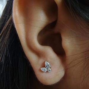 May include: A small, silver butterfly stud earring with three clear, sparkling stones. The earring is set against the skin of an ear, showcasing the delicate design and the way it catches the light. The earring is a piece of jewellery.