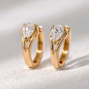 May include: A pair of gold hoop earrings, each featuring a pear-shaped, clear gemstone. The earrings have a polished finish and a modern design. The gemstones are set in a gold bezel setting. The background is a soft, neutral color.