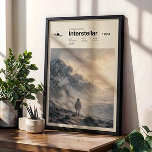 Puede incluir: Un póster de película enmarcado de Interstellar, con fondo beige, texto en negro y una imagen central de un astronauta de pie en un paisaje montañoso nevado bajo un cielo brillante. El póster incluye los créditos de la película y el año 2014.