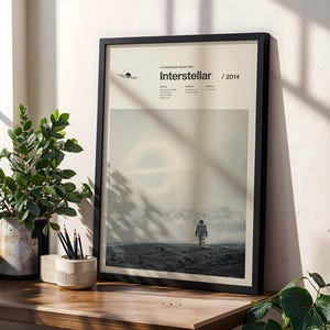 Op de afbeelding: Een ingelijste verticale poster voor de film Interstellar, 2014, met een zwarte lijst, een crèmekleurige achtergrond en een centrale afbeelding van een astronaut die op een donker, rotsachtig landschap naar een gloeiende ring loopt.