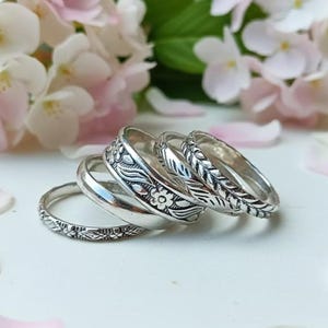 Può includere: Una collezione di anelli d'argento con motivi floreali e a foglie. Gli anelli sono impilati su una superficie bianca, con uno sfondo sfocato di fiori rosa e bianchi e foglie verdi. Gli anelli presentano disegni intricati.