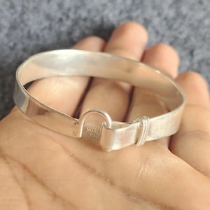 May include: A silver-colored bangle bracelet with a hook clasp. The bracelet is made of a flat, rectangular band of metal that curves around to form a circle. The clasp is a simple hook and loop design.
