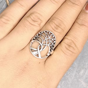 May include: Silver-toned ring featuring a tree of life design. The ring has an oval shape with an openwork pattern depicting the tree's branches and leaves. The ring is worn on a hand with polished nails.