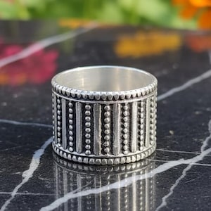 Peut inclure: Une bague en argent avec un design détaillé. La bague présente des lignes verticales de petits points et lignes, créant un motif texturé autour de l'anneau. La bague est présentée sur une surface en marbre noir avec des fleurs colorées en arrière-plan.