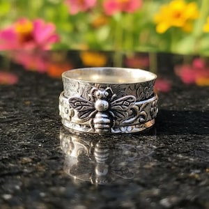 May include: Silver ring with a bee motif. The ring has a textured band with floral and bee patterns. The bee is the central design, with detailed wings and body. The ring is shown on a dark, reflective surface, with a blurred floral background.