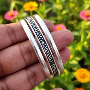 May include: Three silver bangles with a black geometric pattern and a small diamond-like accent. The bangles are held in a hand, with a blurred background of colorful flowers.