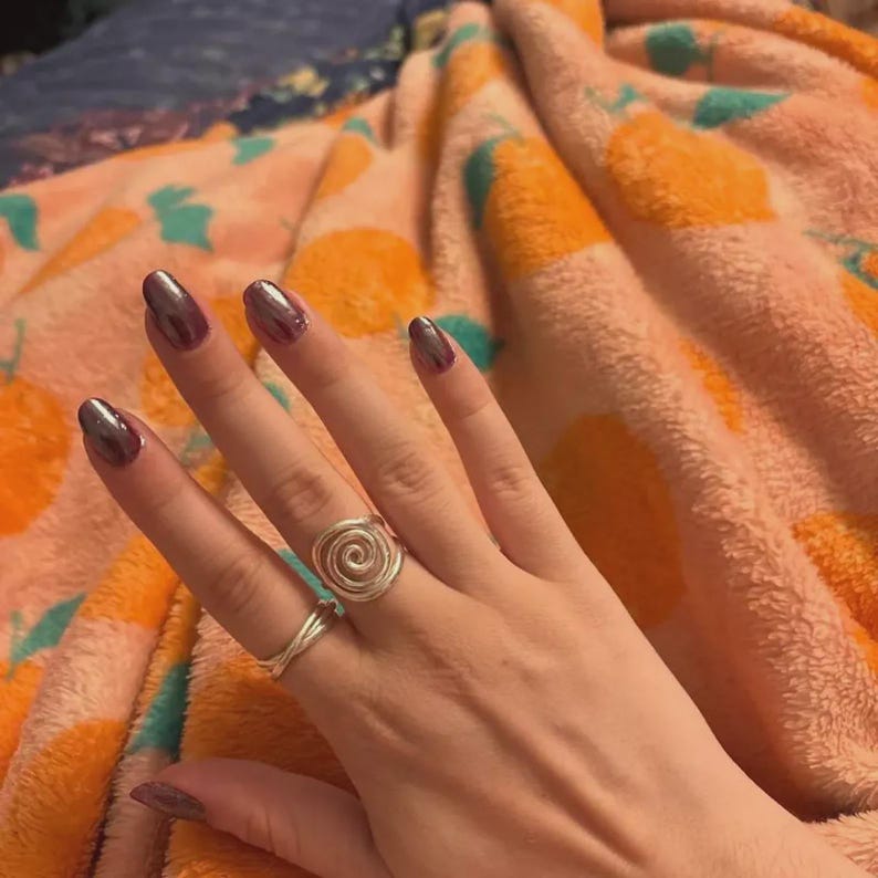 May include: A hand with oval-shaped, metallic purple nails. Two silver rings are worn on the ring finger and middle finger. The ring on the middle finger has a spiral design. The background is a peach-colored blanket with orange and teal patterns.