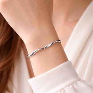 May include: A silver-colored, wavy cuff bracelet. The bracelet has a fluid, organic design, appearing to gently wrap around the wrist. The jewelry is worn on a person's wrist, against a white long-sleeved shirt.