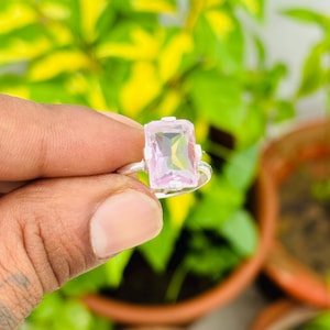 May include: A silver ring featuring a rectangular, light pink gemstone. The gemstone is set in a simple, elegant design. The ring is held up against a background of green foliage.
