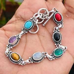 May include: A silver bracelet featuring oval-shaped gemstones in various colors, including red, black, green, blue, yellow, and purple. The bracelet has a toggle clasp and is displayed on a hand.