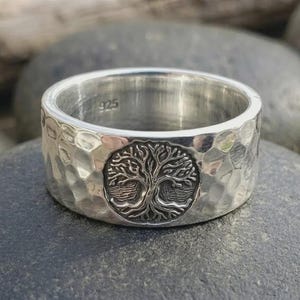 May include: A wide sterling silver ring featuring a hammered, textured finish on the outer band. The center of the band displays a detailed, circular embossed design of a tree with spreading branches and roots. The interior is stamped with 925.