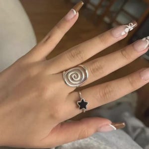 May include: A hand wearing two silver rings. One ring features a spiral design, and the other has a black star. The nails are painted with a brown and white design.