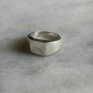 May include: A silver signet ring with a sunburst design engraved on the face. The ring has a rectangular face and a thick band. The metal is polished and reflects light. The ring is a size 8.5.